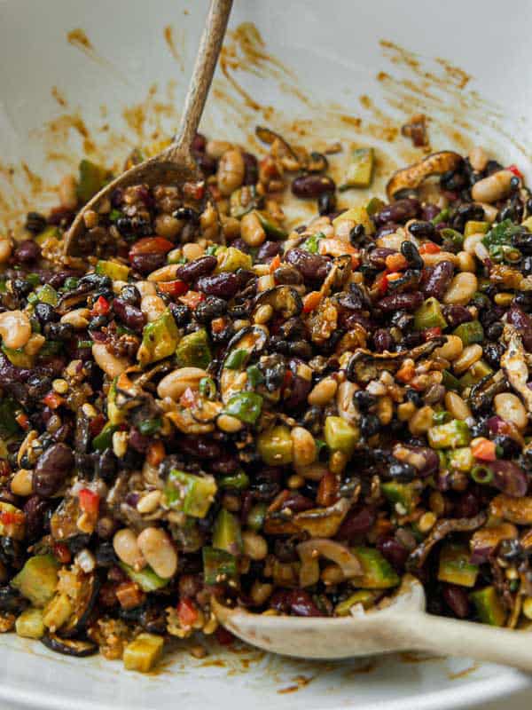 A close-up of a colorful dense bean salad with diced vegetables, black beans, kidney beans, pinto beans, corn, red peppers, and herbs, mixed together in a white bowl with two wooden serving spoons.